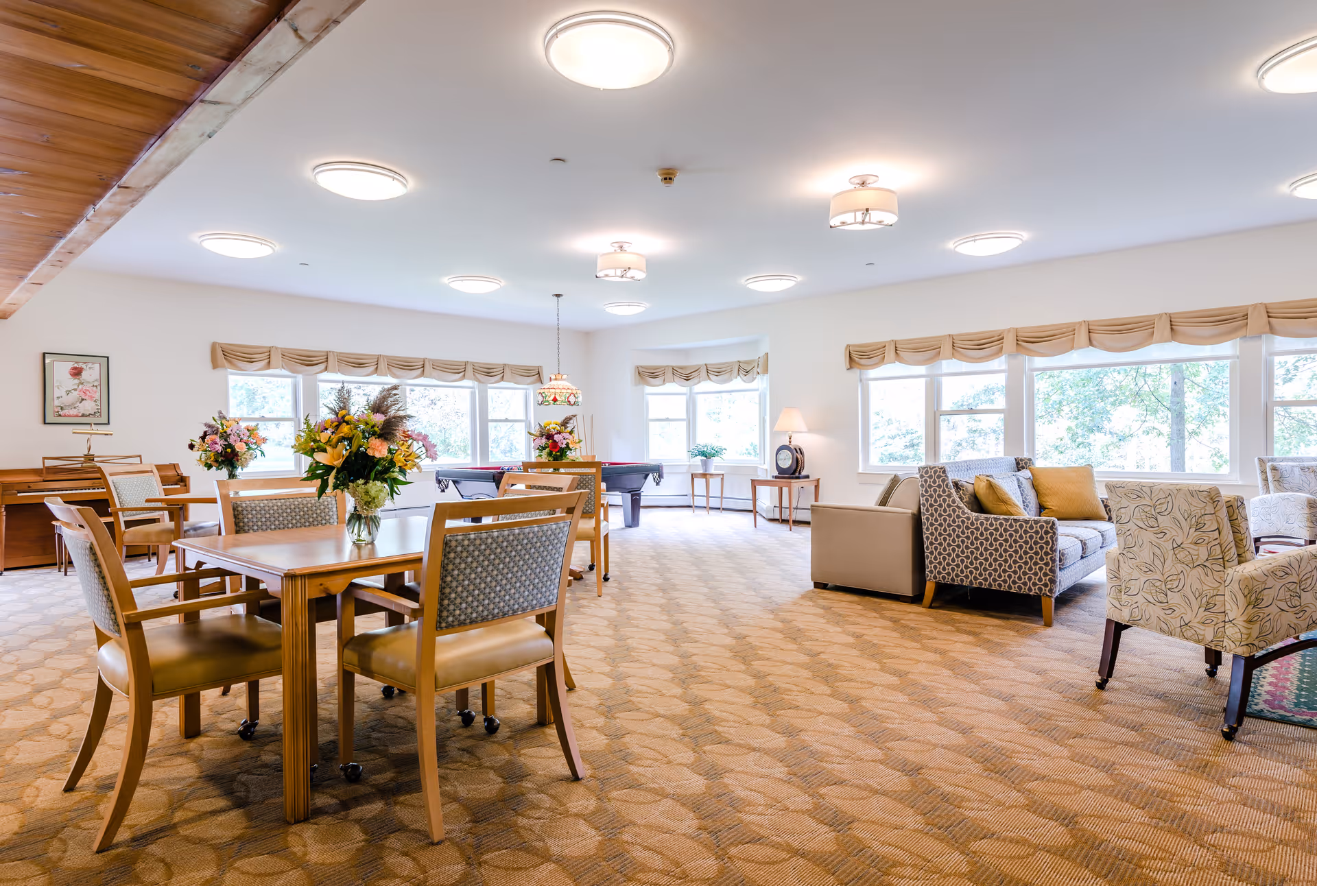 Bright communal lounge and dining area with wooden tables and chairs, sofas, and large windows.