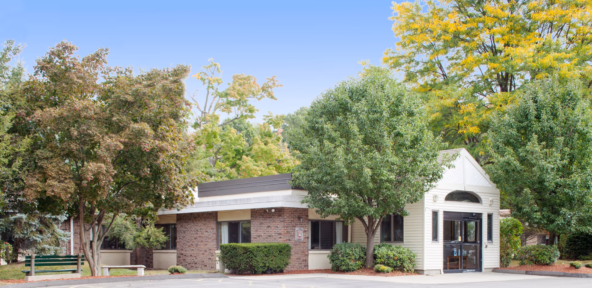 Exterior view of a single-story building with a combination of brick and white siding, surrounded by green trees and bushes under a clear blue sky. There is a parking area in front and a green bench on the left side near the trees.