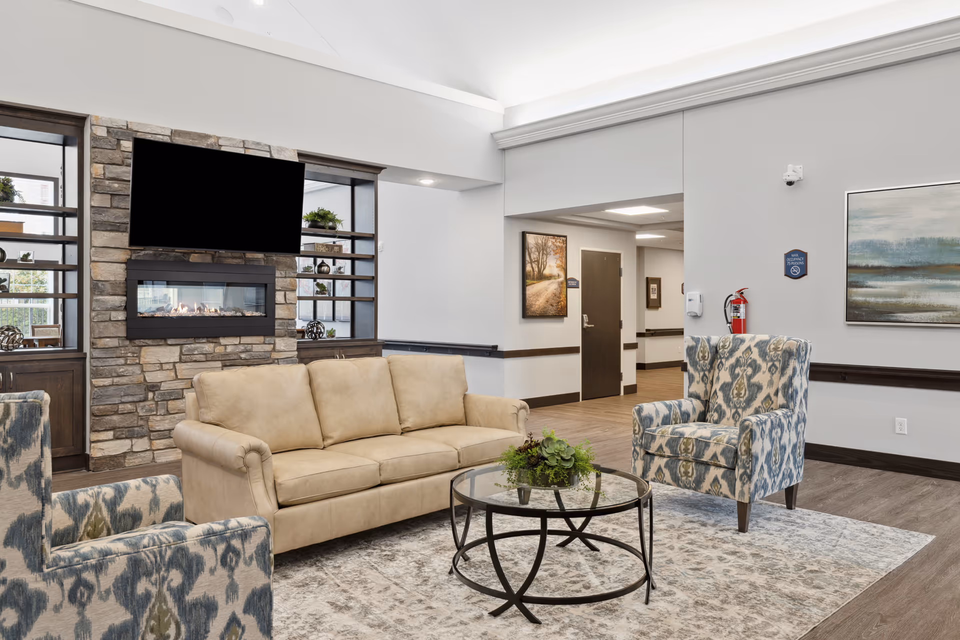 A cozy senior living facility common area with a beige sofa and two patterned armchairs arranged around a round glass coffee table with a plant centerpiece. Behind the sofa is a stone fireplace with a mounted flat-screen TV above it. The room has light-colored walls, wood flooring, and framed artwork on the walls.