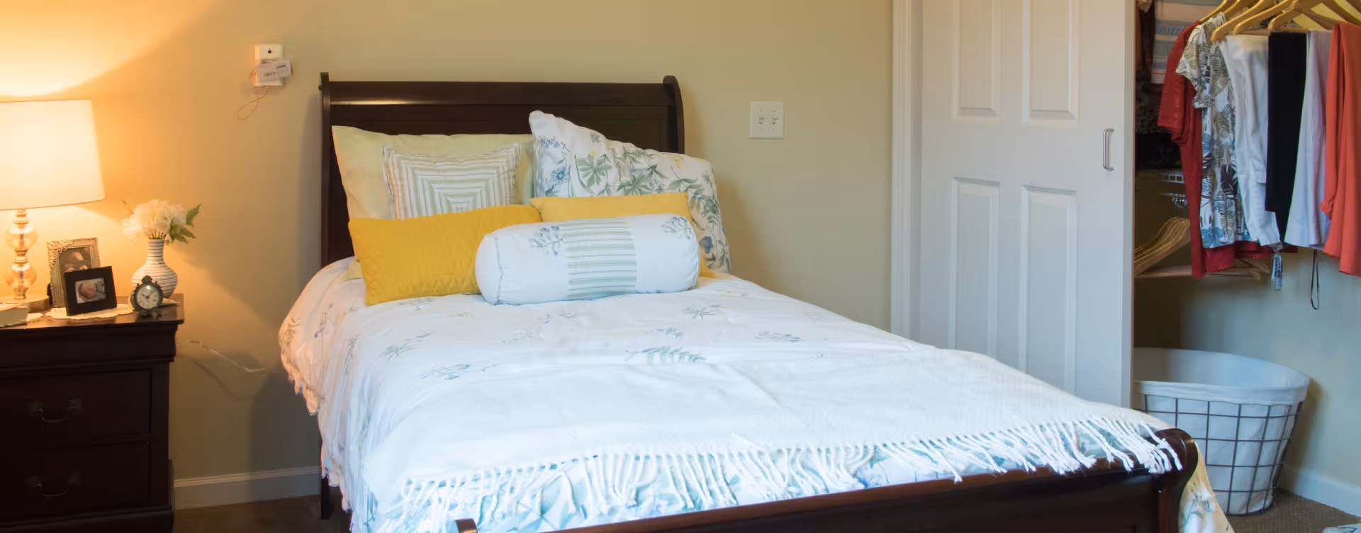 A cozy bedroom with a wooden bed frame, a bed made with white and floral bedding, and several pillows including a yellow one. To the left of the bed is a dark wooden nightstand with a lamp, a small clock, a framed photo, and a vase with flowers. On the right side, there is an open closet with hanging clothes and a laundry basket on the floor.