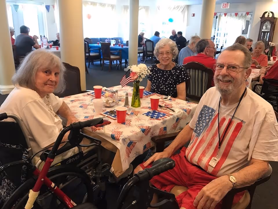 Three senior residents sit around a table decorated with a patriotic tablecloth, flags, and cups in a communal dining room.