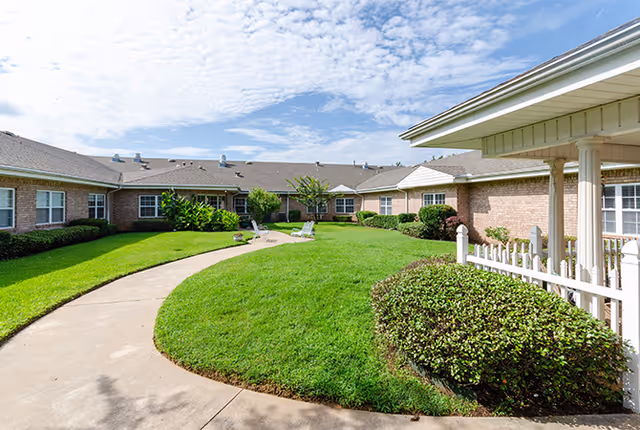Outdoor courtyard area of a senior living facility with a curved concrete walkway, well-maintained green grass, shrubs, and brick buildings surrounding the space under a partly cloudy sky.