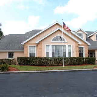 Front exterior view of a single-story building with peach-colored walls, white trim, and a gray shingled roof. There is a large window with an arched top in the center, flanked by smaller windows. An American flag is mounted on a pole in front of the building, and neatly trimmed bushes line the front. The sky is partly cloudy.
