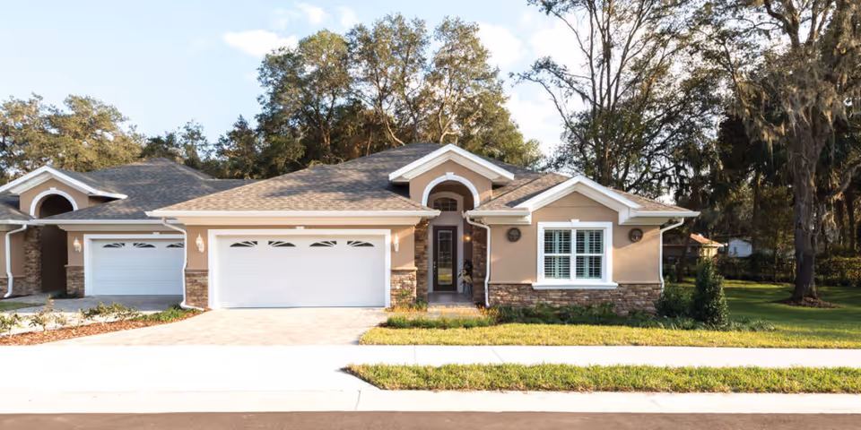 Exterior front view of a single-story residential building with a two-car garage, beige walls, stone accents, and a shingled roof, surrounded by grass and trees under a partly cloudy sky.