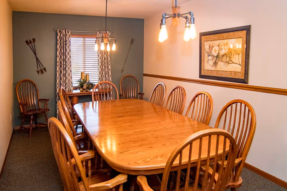 Long wooden dining table surrounded by high-back wooden chairs in a warmly lit dining room with a window and wall art.