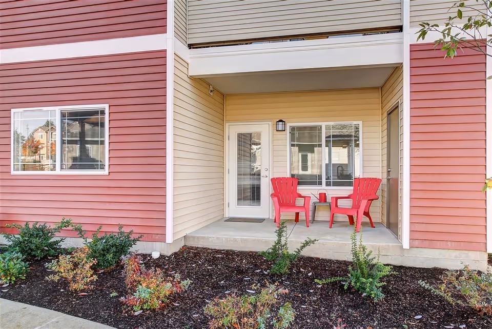 Outdoor patio area of a residential building with beige and red siding. The patio has two red Adirondack chairs and a small table between them. There are plants and shrubs in the garden bed in front of the patio.