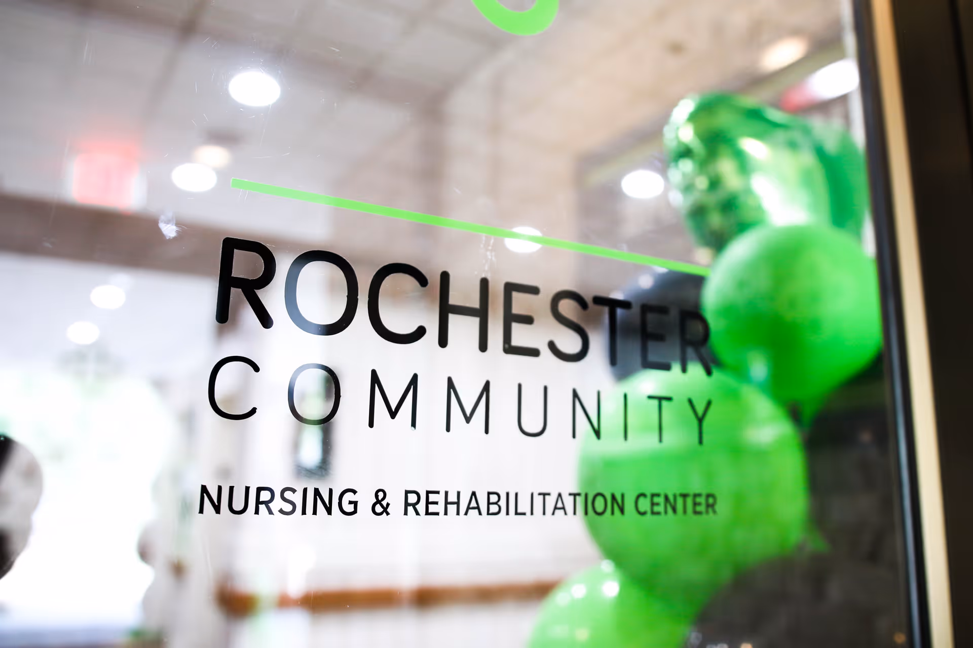 Glass entrance door reading "Rochester Community Nursing & Rehabilitation Center" with green balloons visible inside.