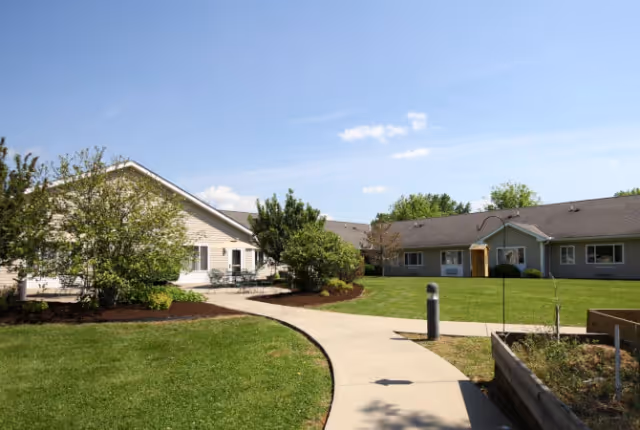 Outdoor courtyard area of a senior living facility with a curved concrete walkway, green grass, bushes, and trees. Single-story buildings with beige and gray siding surround the courtyard under a clear blue sky.