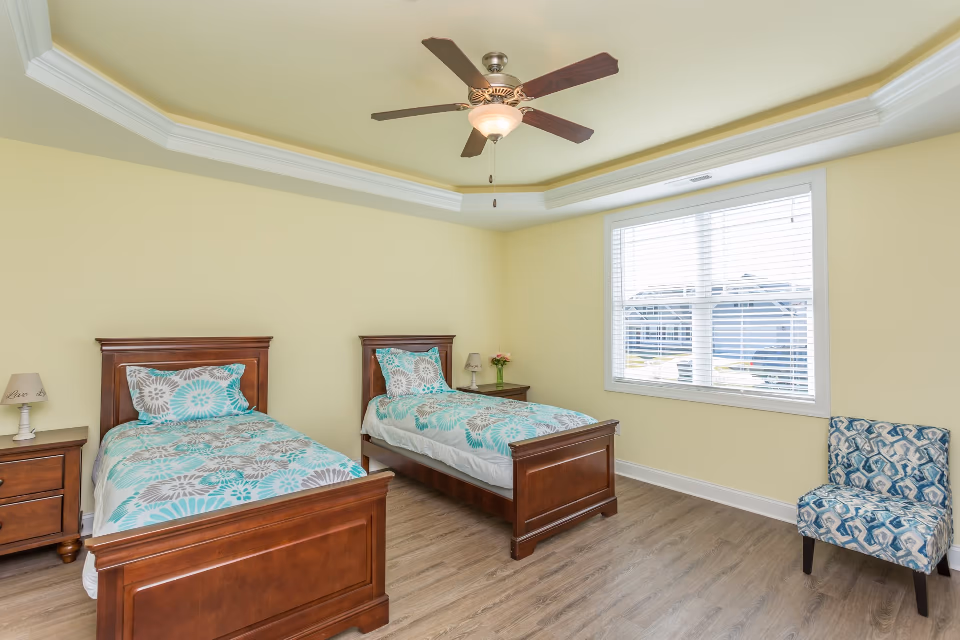 Bright bedroom with two wooden twin beds with teal-patterned bedding, a ceiling fan, nightstands, and a window.