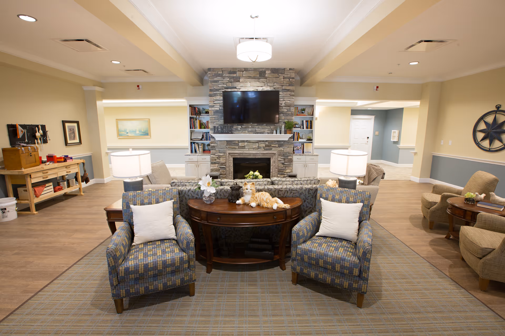 A cozy living room area in a senior living facility with two patterned armchairs with white pillows facing a stone fireplace with a mounted flat-screen TV above it. Behind the armchairs is a wooden console table with a flower vase and a stuffed cat toy. The room has warm lighting, wooden flooring, and additional seating areas with armchairs and side tables. Bookshelves flank the fireplace, and the walls are painted in soft yellow and blue tones.