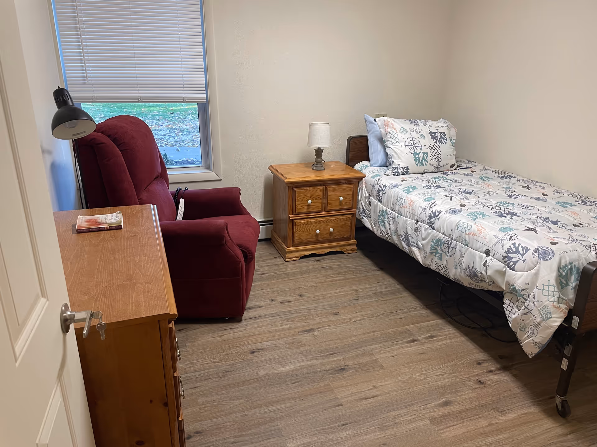 A small, simple bedroom with a single bed covered in a patterned comforter, a wooden nightstand with a lamp, a red upholstered recliner chair, a wooden dresser with a book on top, and a window with closed blinds.