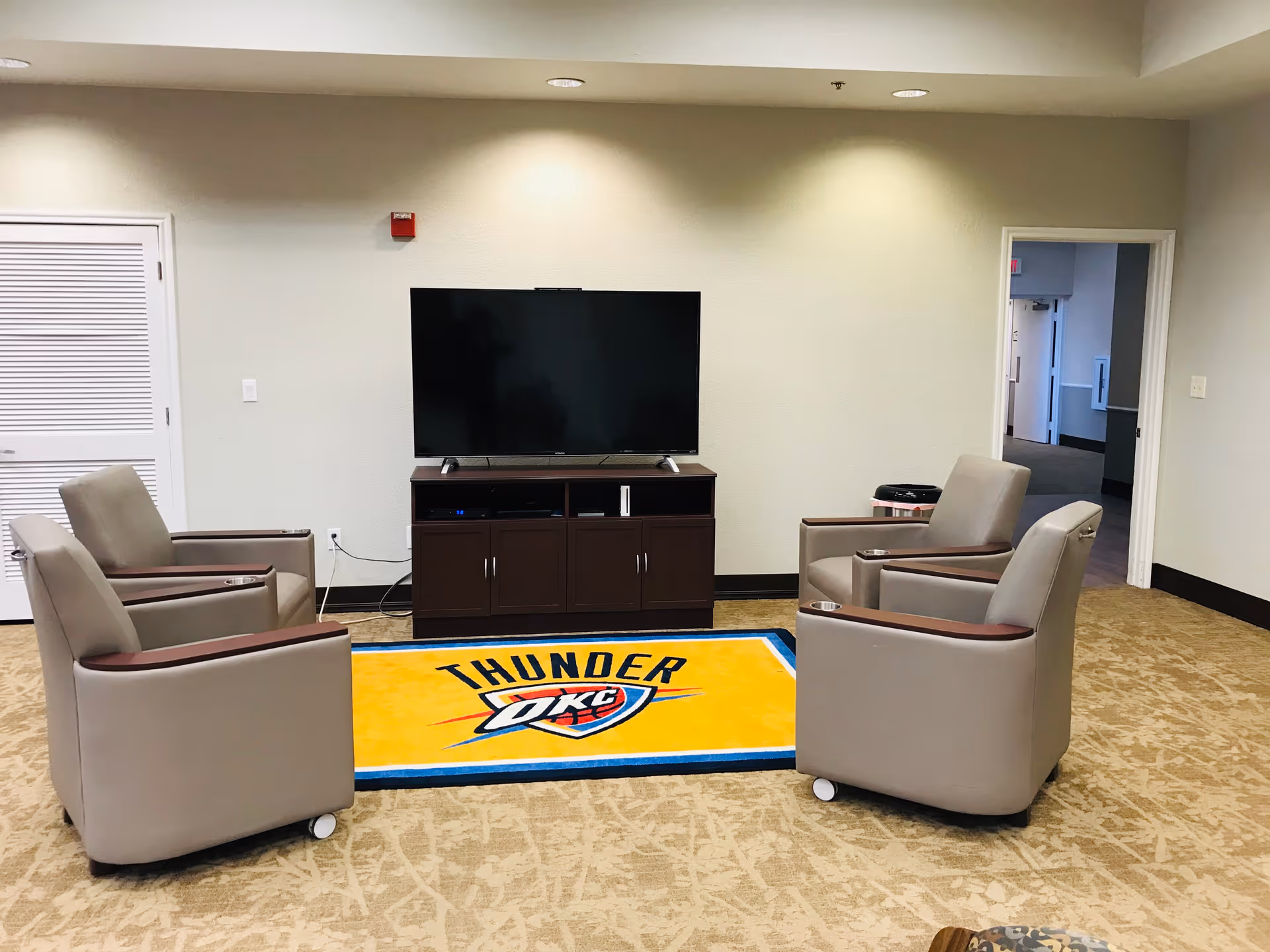 A common area with four beige armchairs arranged around a TV on a dark wooden stand. A yellow rug with the Oklahoma City Thunder basketball team logo is placed in front of the TV. The room has beige walls and carpet, with a doorway leading to another room on the right side.