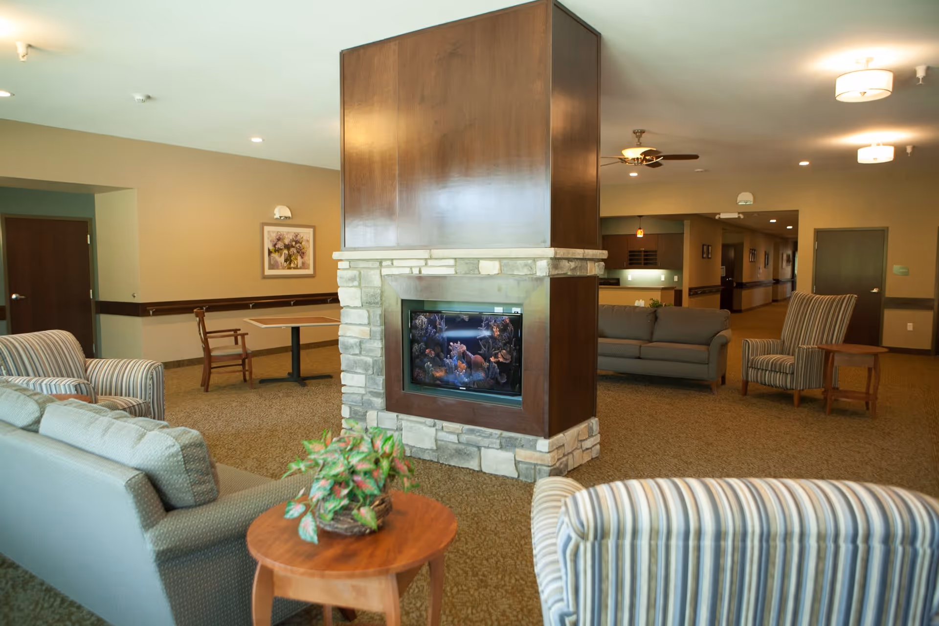 A cozy assisted living common area featuring a central stone and wood fireplace with an aquarium inside. Surrounding the fireplace are comfortable sofas and striped armchairs, a small wooden table with a potted plant, and a table with chairs against the wall. The room has beige walls, carpeted floors, ceiling lights, and a ceiling fan.