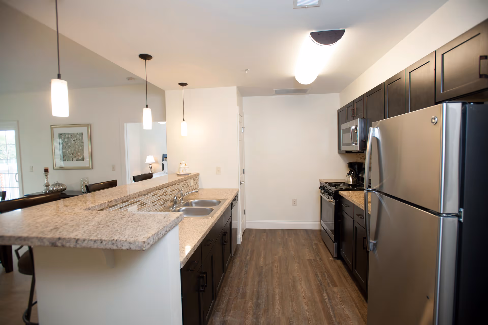 Modern kitchen with granite countertops, dark wood cabinets, stainless steel refrigerator, oven, and microwave. The kitchen has a double sink and pendant lights hanging over the counter. The floor is wood-style and the kitchen opens to a dining area with a framed picture on the wall.