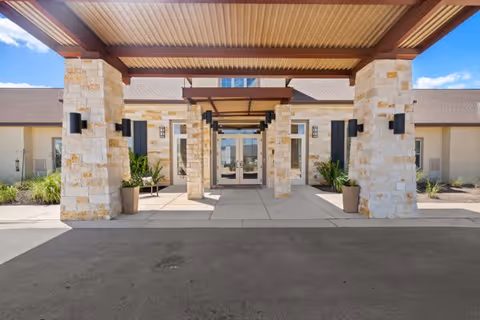 Entrance of a senior living facility with a covered driveway supported by stone pillars, glass double doors, and potted plants on either side under a clear blue sky.