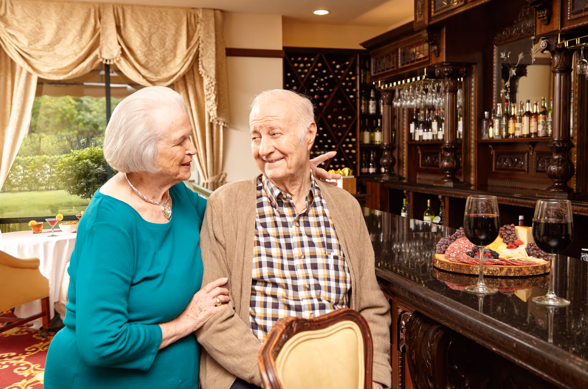 An elderly couple smiling and sitting together at a dark wooden bar counter in a warmly lit room. The bar is stocked with bottles and glasses, and there is a platter with cheese, grapes, and salami along with two glasses of red wine on the counter. Behind them, there is a window with beige curtains showing a green outdoor garden area.