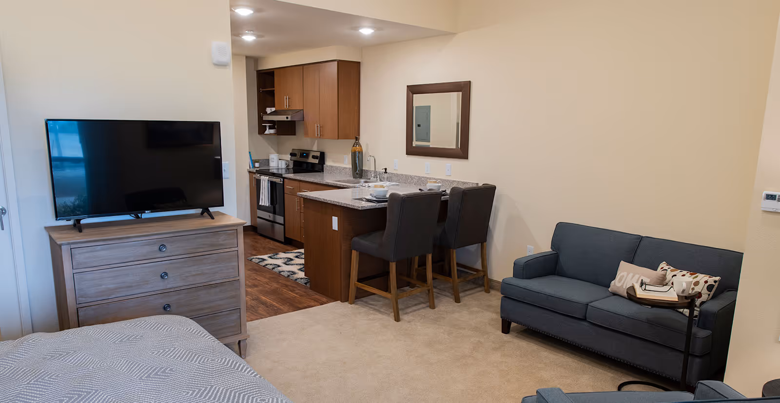 Interior view of a senior living facility apartment at Westgate Village featuring a small kitchen with wooden cabinets, a granite countertop with two bar stools, a gray sofa with decorative pillows, a TV on a wooden dresser, and part of a bed with a patterned gray bedspread.