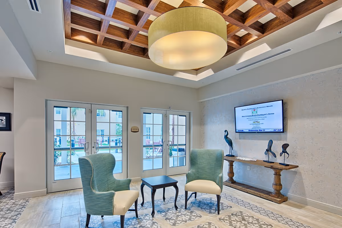 A bright and airy common area with two green upholstered armchairs and a small black table between them. The room features a decorative wooden coffered ceiling with a large round light fixture. There are double glass doors leading outside, and a wooden console table against the wall with three bird sculptures. A flat-screen TV is mounted on the wall above the console table.