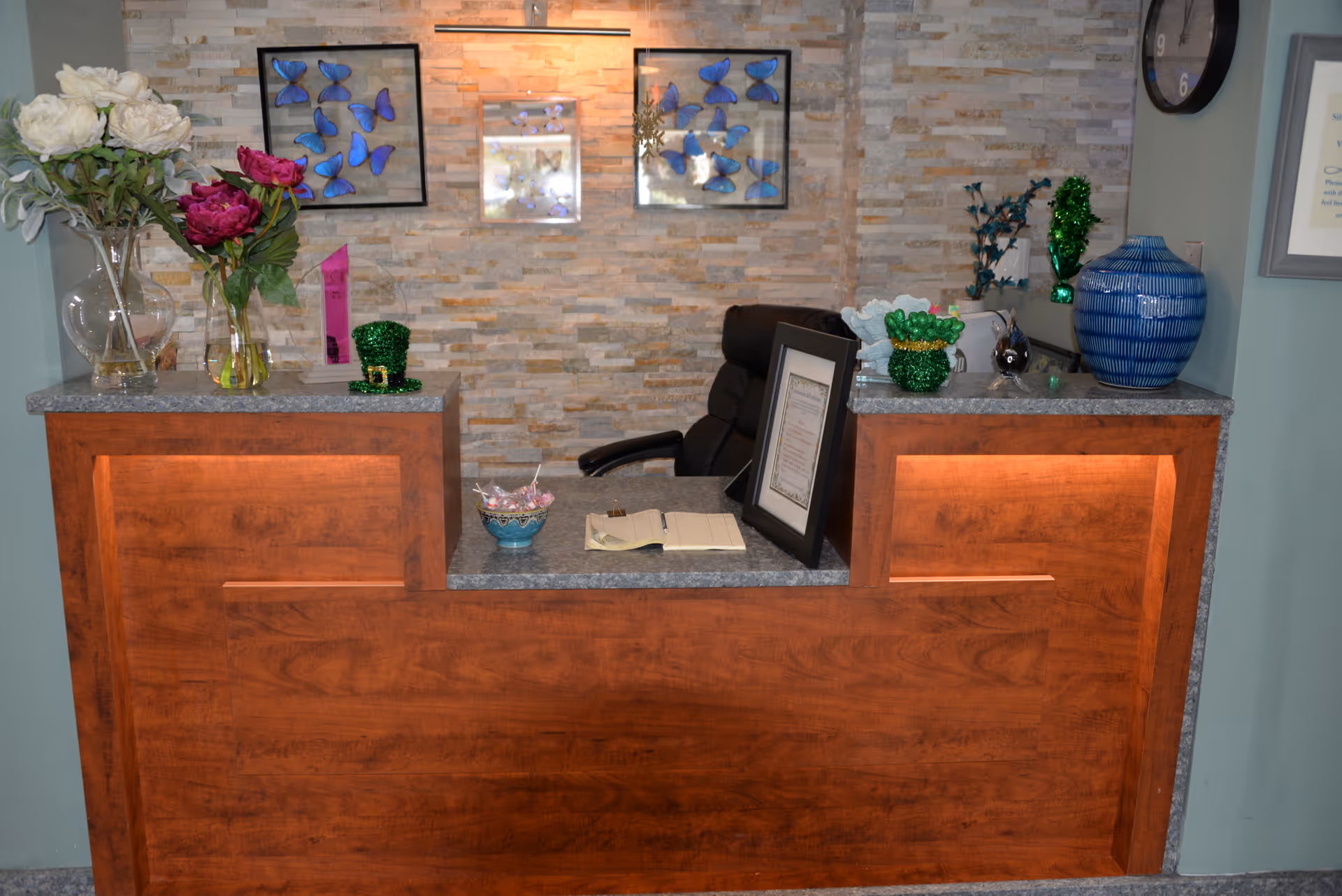 Reception desk area with a wooden counter and granite top, decorated with vases of flowers, framed butterfly artwork on a stone accent wall, a black office chair behind the desk, and various decorative items including a green glittery hat and a blue ceramic vase.