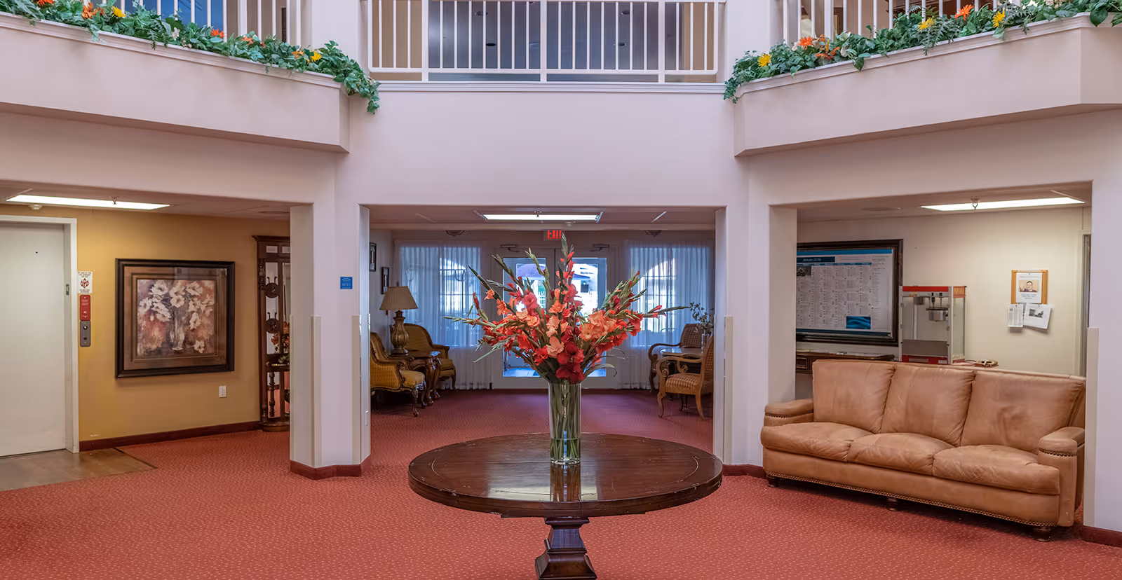 Entrance lobby with a round table holding a vase of red flowers, seating areas, and an overhead balcony with planter boxes.