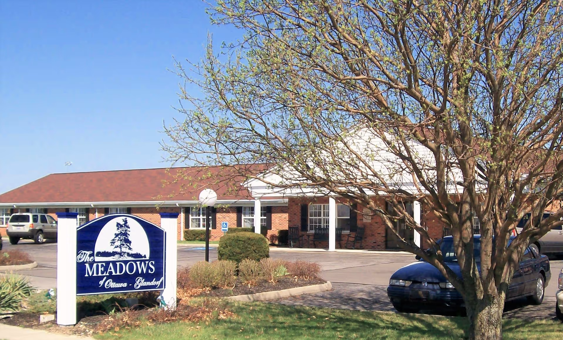 Exterior view of The Meadows of Ottawa senior living facility showing a brick building with a red roof, a parking lot with several cars, a tree with budding leaves in the foreground, and a blue and white sign with the facility name.