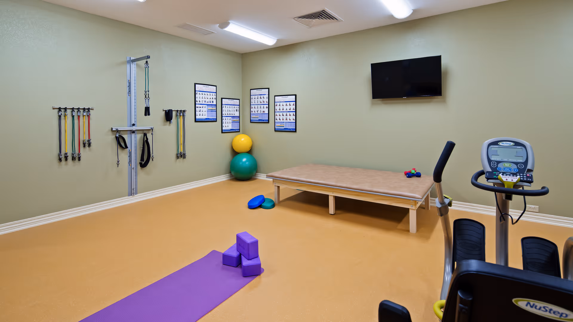 Bright therapy and exercise room with a yoga mat and blocks, resistance bands on the wall, exercise balls, a treatment table, and cardio equipment.