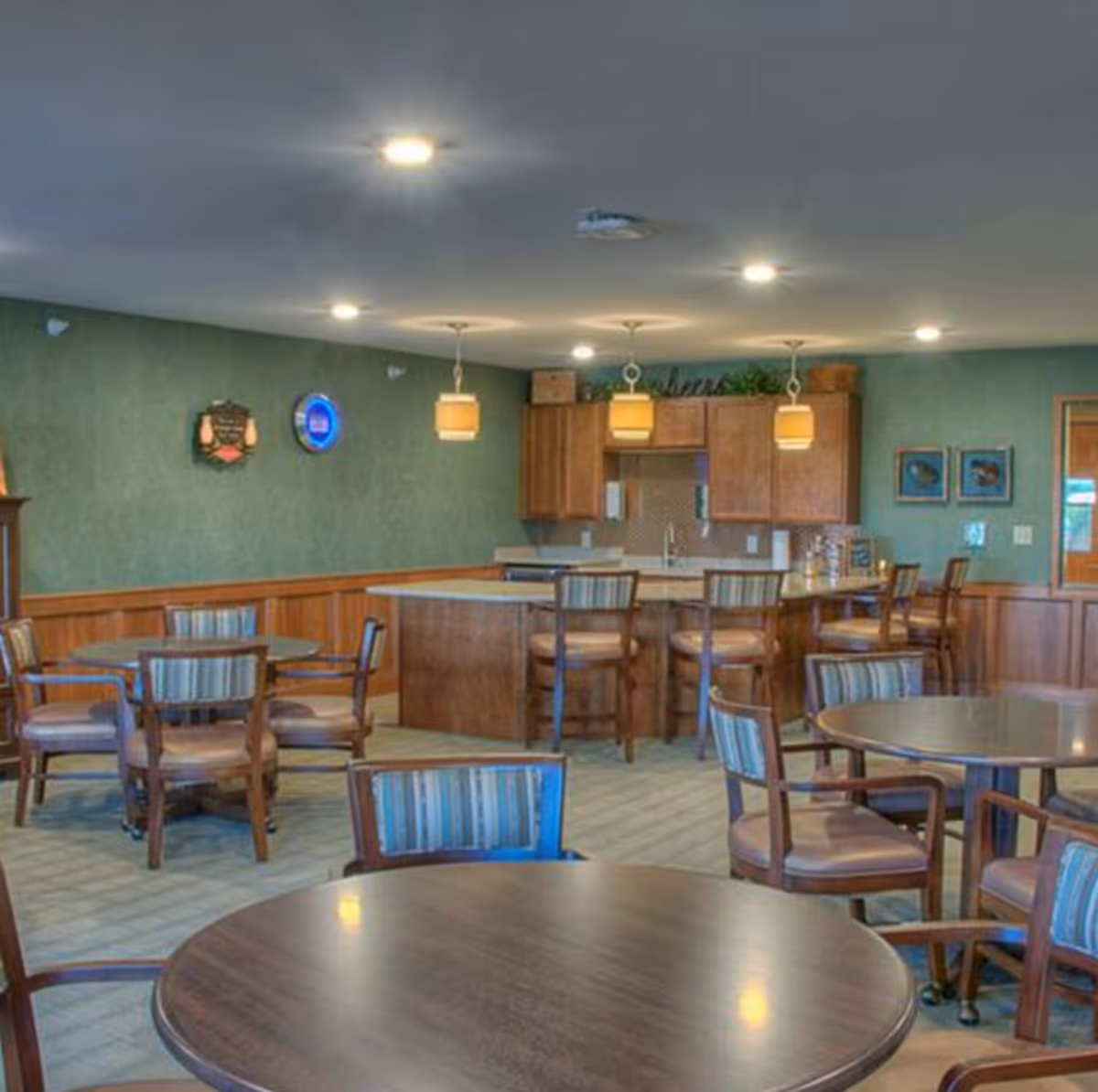 Interior view of a communal dining area with round tables and wooden chairs featuring striped cushions. In the background, there is a kitchen area with wooden cabinets, a countertop with bar stools, and pendant lights hanging from the ceiling. The walls are green with wooden paneling on the lower half, and there are decorative items and framed pictures on the walls.