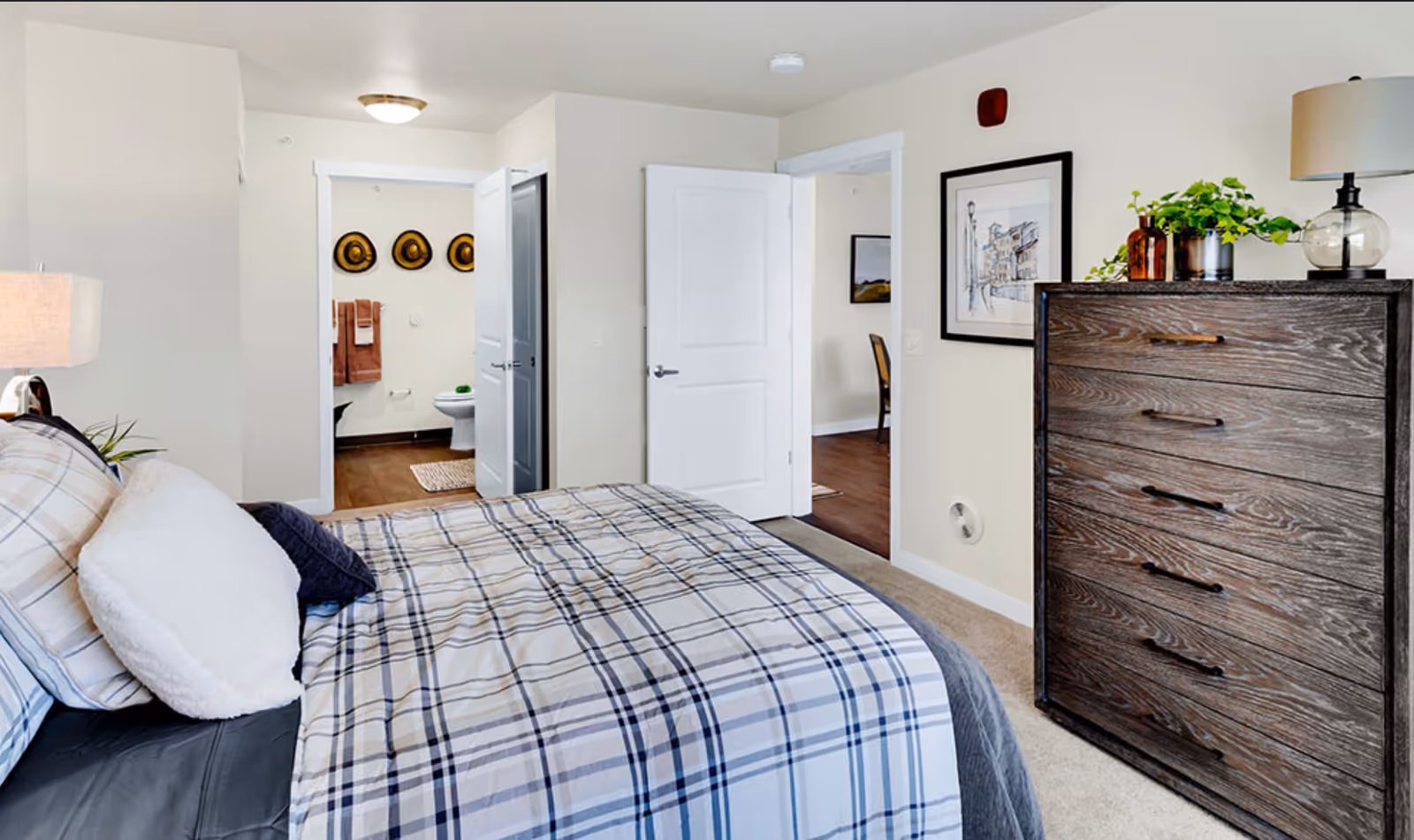 A bedroom with a bed covered in a plaid comforter and multiple pillows. To the right is a dark wooden dresser with a lamp, plants, and decorative items on top. The room has beige walls and carpeted floor. Two open doors reveal a bathroom with three decorative hats on the wall and a toilet, and another room with a wooden floor and a chair.