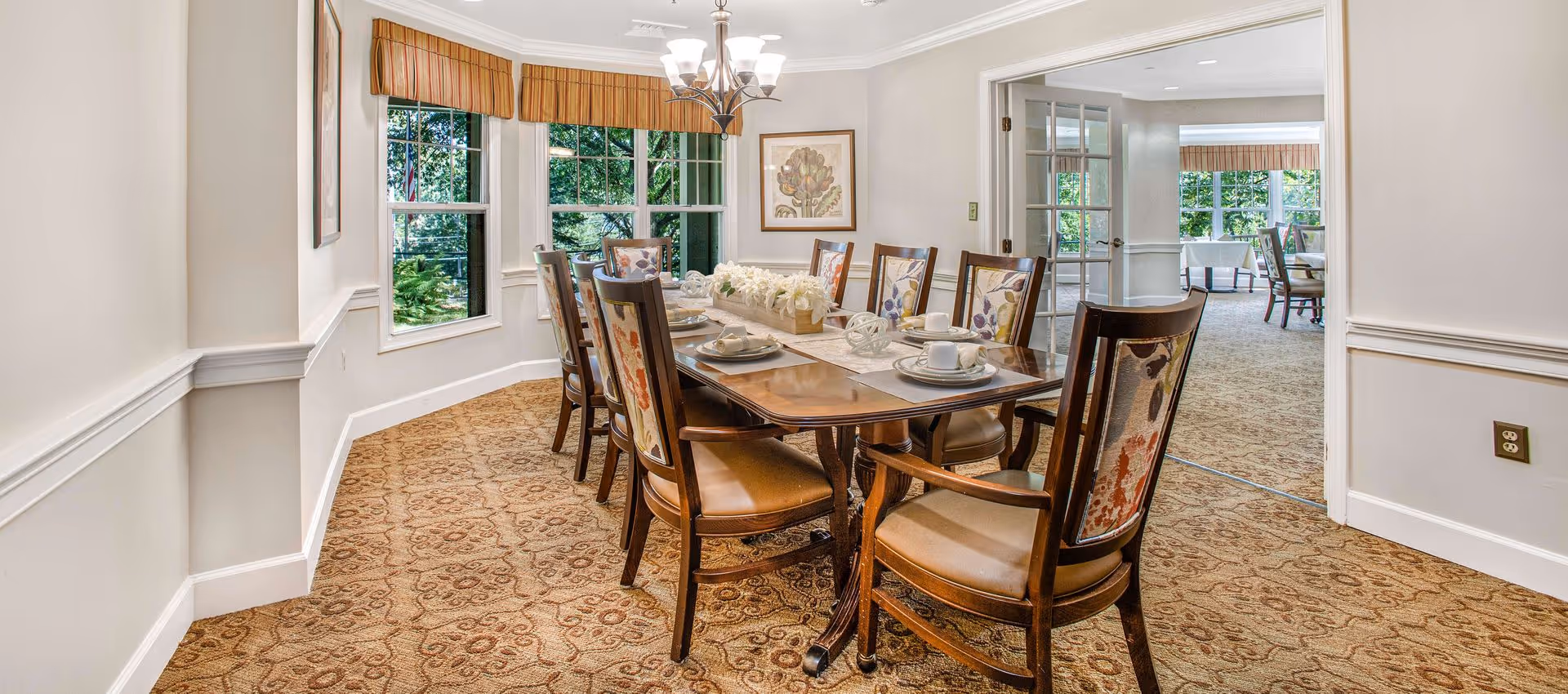 A well-lit dining room with a long wooden table set for eight people. The chairs have floral upholstery on the backrests. The room has large windows with striped valances, beige walls, and patterned carpet. A chandelier hangs above the table, and there is a framed botanical print on the wall. French doors open to another room with additional dining tables and chairs.
