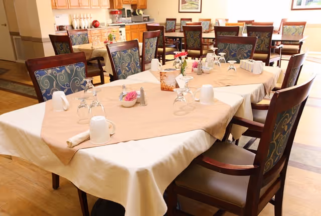 Dining room with tables covered in beige and white tablecloths, set with upside-down glasses, cups, napkins, and small flower arrangements. Wooden chairs with patterned upholstery surround the tables. Kitchen cabinets and countertops are visible in the background.