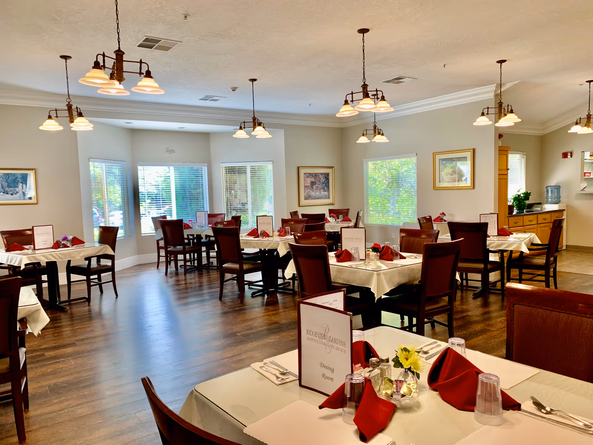 A bright and spacious dining room with multiple tables covered in white tablecloths and set with red napkins, silverware, and glasses. The room has wooden floors, several windows letting in natural light, framed artwork on the walls, and multiple hanging light fixtures. A sign on one table reads 'Ridgeview Gardens Assisted Living Dining Room.'