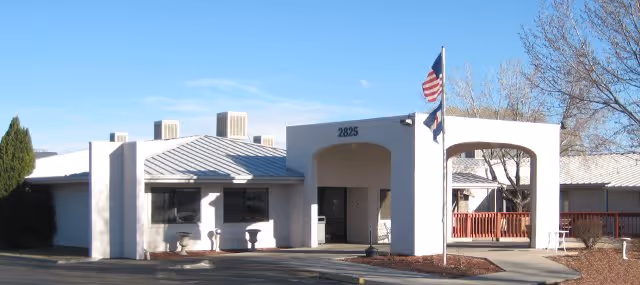 Exterior view of a single-story building with a white facade and a metal roof, featuring an entrance with an archway and two flagpoles displaying the American flag and another flag. The building number 2825 is visible above the entrance. There are some trees and shrubs around the building under a clear blue sky.