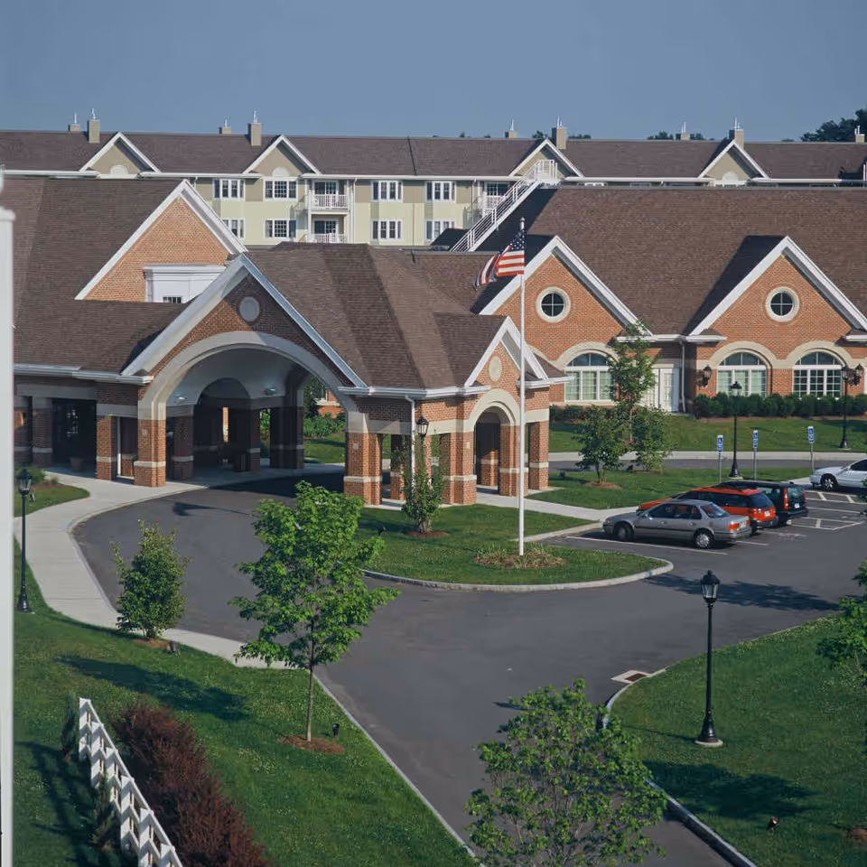 Exterior front entrance of a brick senior living building with a covered portico, American flag, parked cars, and landscaped grounds.