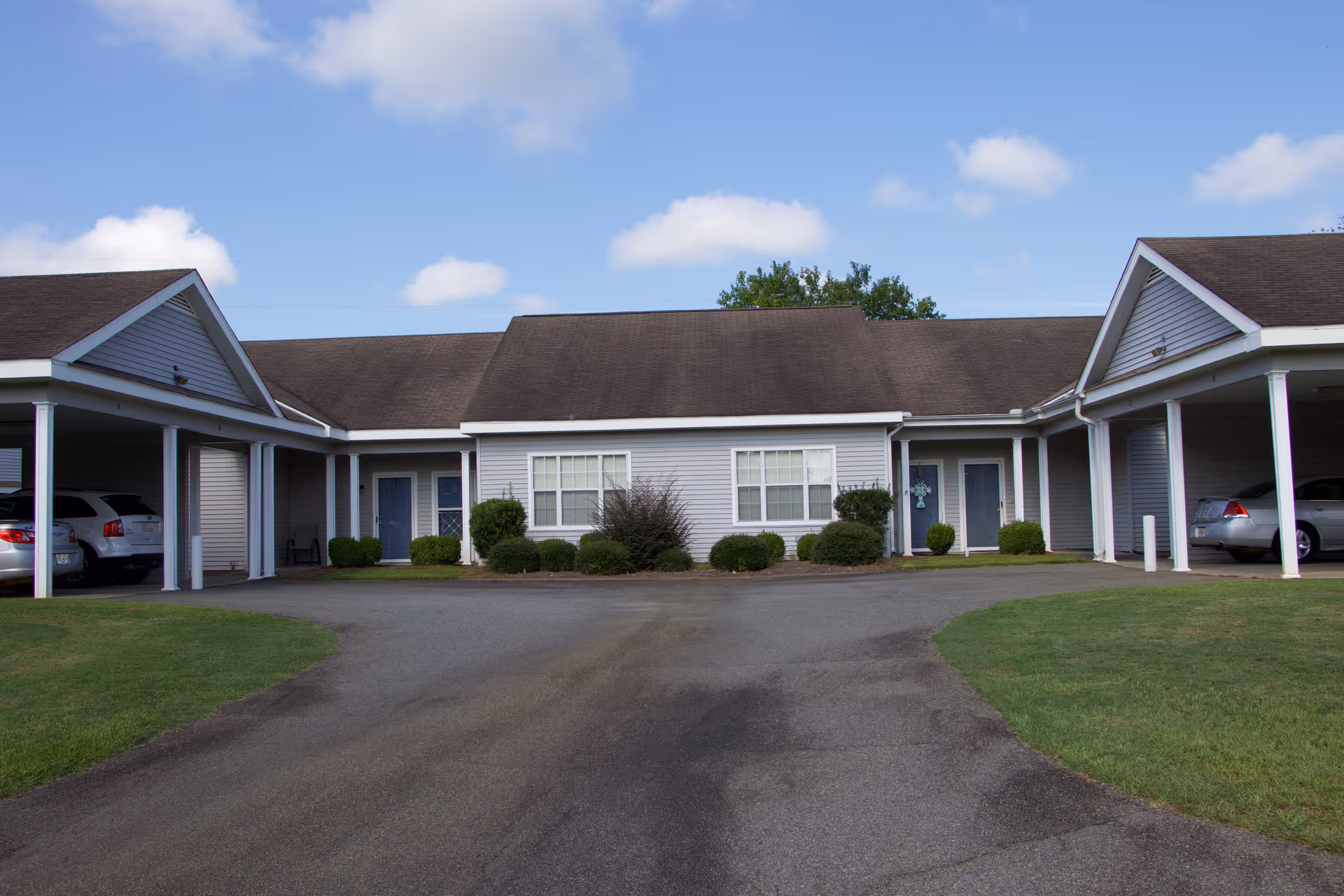 Exterior view of Magnolia Manor of Americus showing a single-story building with gray siding, white trim, and a brown shingled roof. The building has two covered carports on either side with parked cars, a paved driveway, green grass, and some bushes in front of the windows. The sky is partly cloudy.