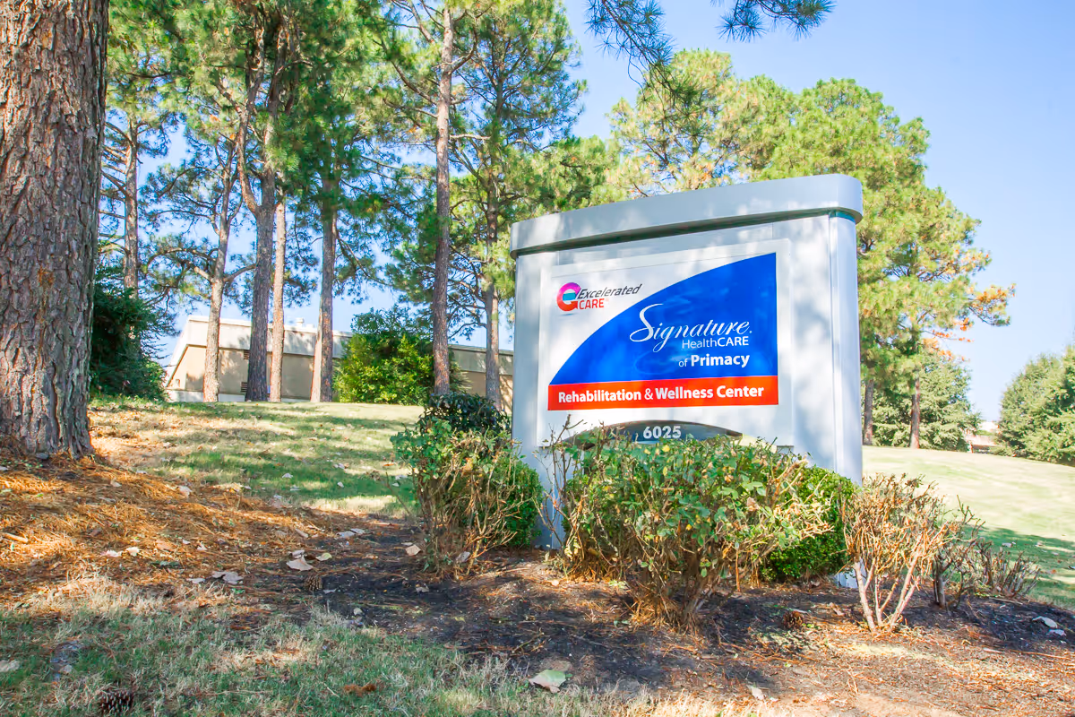 Sign for Signature HealthCARE of Primacy Rehabilitation & Wellness Center on a landscaped lawn with trees and a building in the background.