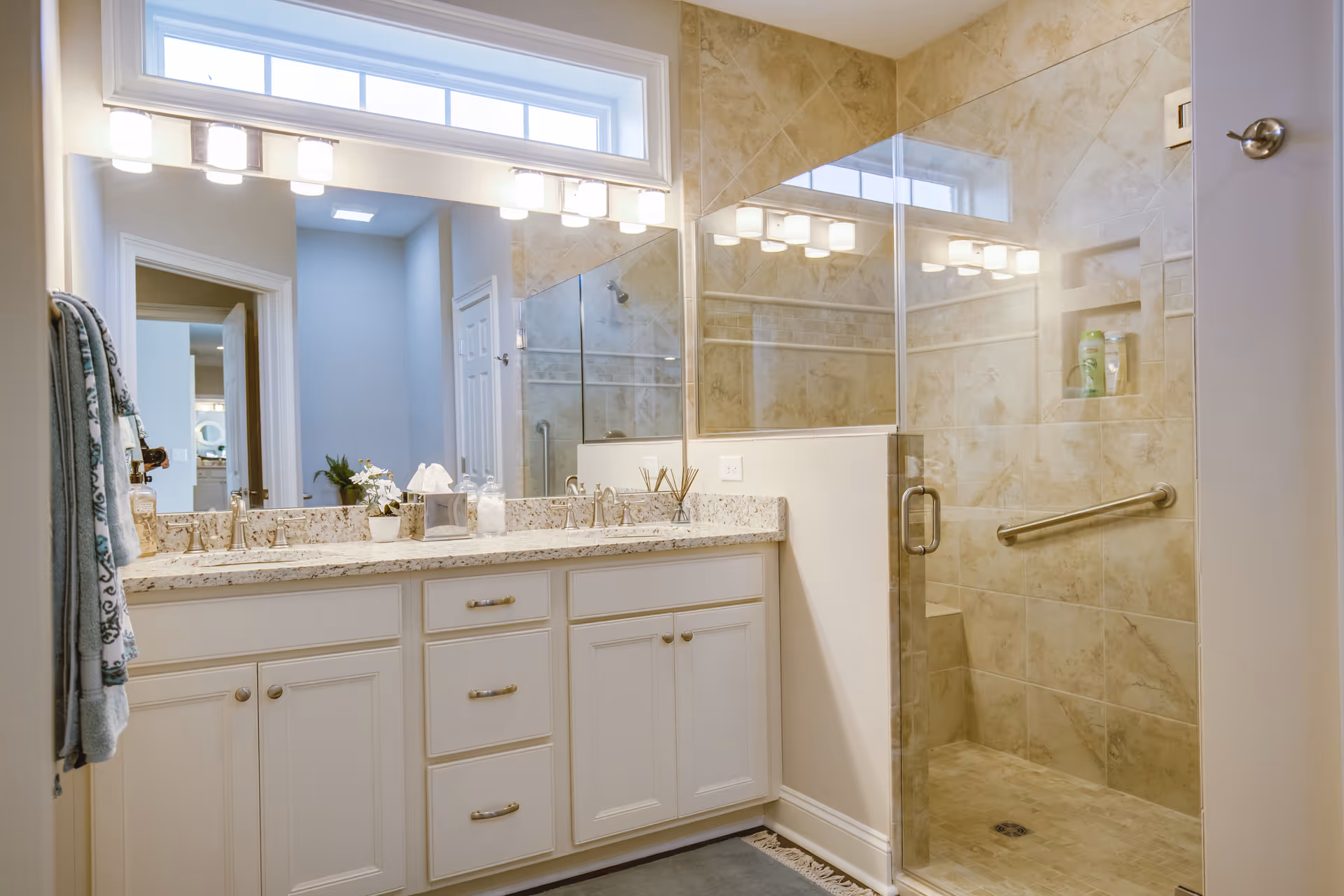 A clean and modern bathroom featuring a large vanity with a granite countertop, two sinks, and multiple drawers and cabinets. Above the vanity is a large mirror with a row of lights and a small window above it. To the right is a walk-in shower with glass doors, beige tiled walls, a built-in shelf with toiletries, and a safety grab bar. Towels hang on a rack to the left side of the vanity.