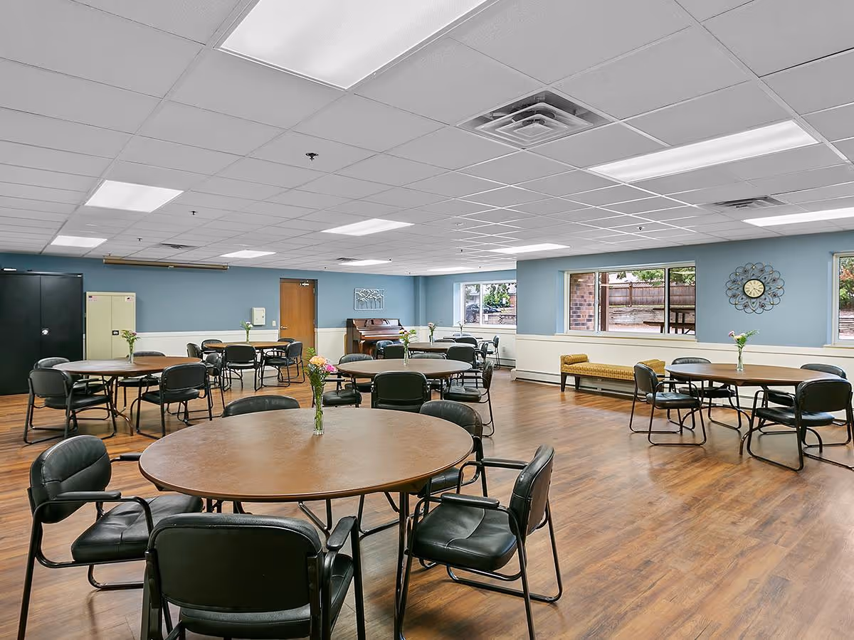 A spacious dining room with multiple round tables surrounded by black chairs. Each table has a small vase with flowers. The room has light blue walls, large windows letting in natural light, a wooden floor, and a piano against the far wall. There is a decorative clock on one wall and a bench beneath the windows.