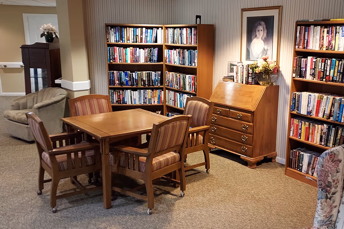 A cozy reading and sitting area in a senior living facility featuring a wooden table with four striped cushioned chairs on wheels, surrounded by bookshelves filled with books. There is a wooden chest of drawers with a flower vase and framed portrait above it, and a cushioned armchair in the background.