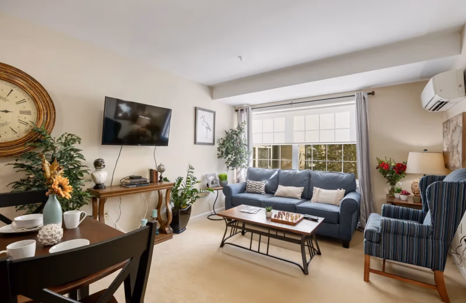 A cozy living room with a blue sofa and striped armchair, a wooden coffee table with a chessboard, a wall-mounted TV above a wooden console table, large window with light curtains, and various plants and decorative items.