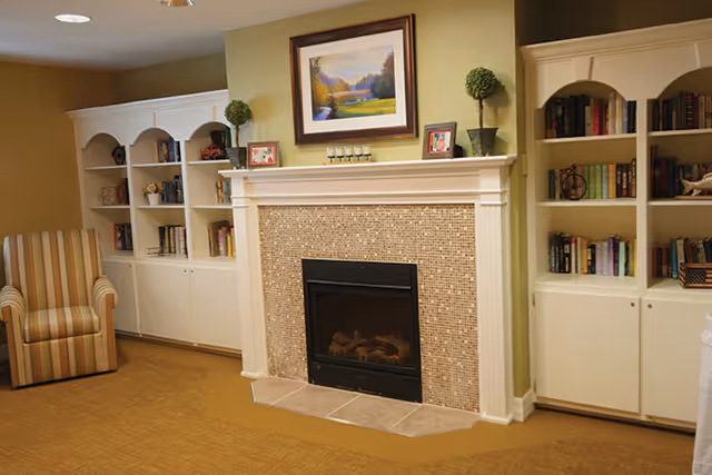 A cozy living room area featuring a fireplace with a mosaic tile surround and white mantel. Above the mantel is a framed landscape painting. On either side of the fireplace are white built-in bookshelves filled with books and decorative items. A striped armchair is positioned to the left of the fireplace. The walls are painted a warm beige color and the floor is carpeted.
