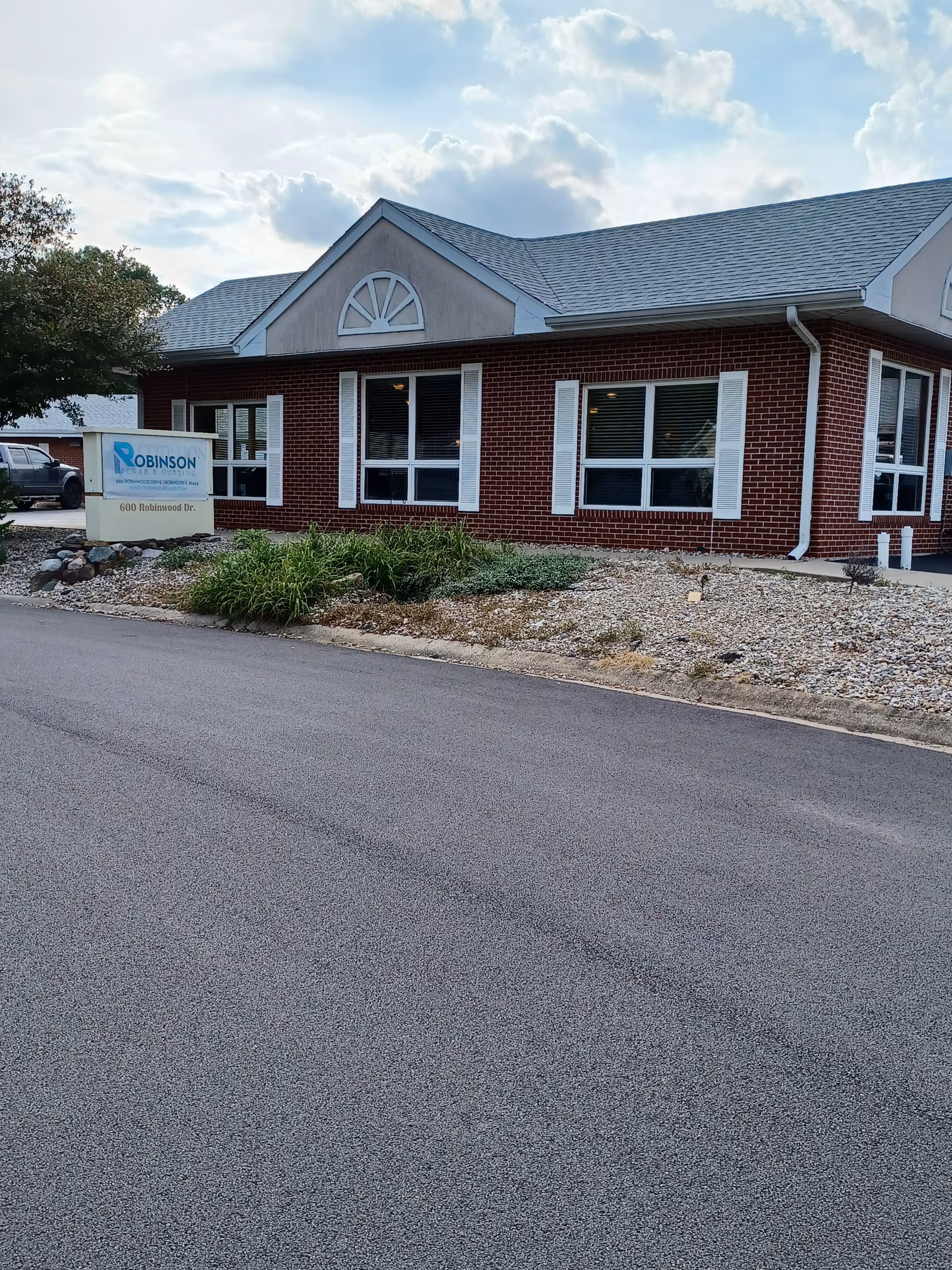 Exterior view of a single-story brick building with white window shutters and a gray shingled roof under a partly cloudy sky. There is a sign in front of the building that reads Robinson Rehab and Nursing LLC, located at 600 Robinwood Dr. The area around the building has landscaping with rocks and some greenery.