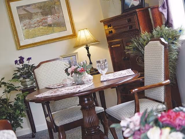 A cozy dining area with a small wooden oval table set for two, featuring placemats, a glass of water, and a floral centerpiece. Two upholstered wooden chairs are positioned around the table. In the background, there is a wooden cabinet, a table lamp, a framed painting on the wall, and several potted plants adding greenery to the room.