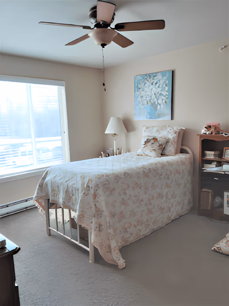 A bedroom with a single bed covered in a floral bedspread and matching pillows. There is a ceiling fan above, a window with blinds letting in natural light, a bedside table with a lamp, and a wooden shelving unit with various items on it. A framed painting of a tree hangs on the wall above the bed.