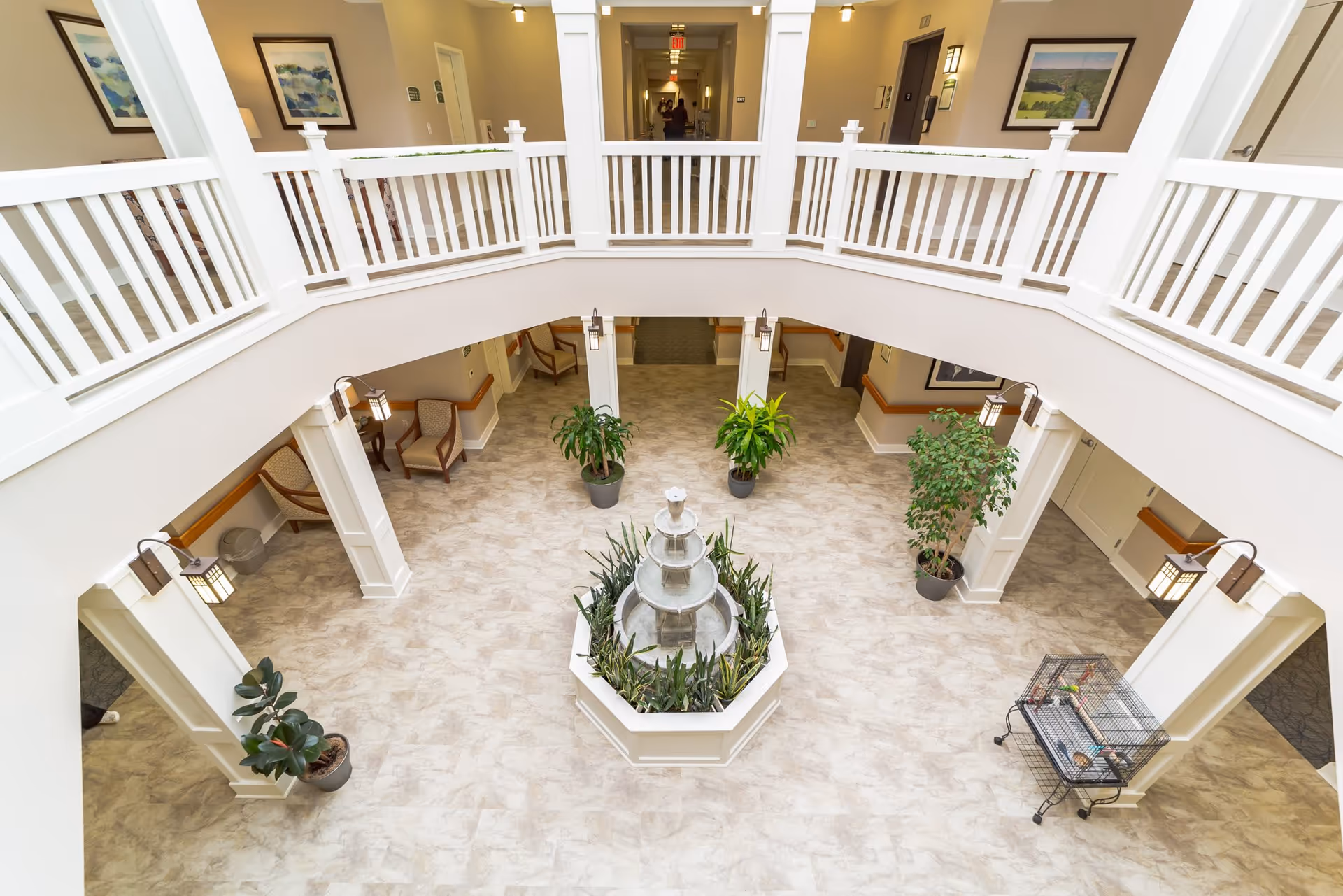 Interior view of a senior living facility atrium with a central three-tiered water fountain surrounded by plants. The space has beige tiled flooring, white railings on the upper level, several potted plants, and chairs along the walls. The area is well-lit with wall-mounted lantern-style lights and framed artwork on the walls.