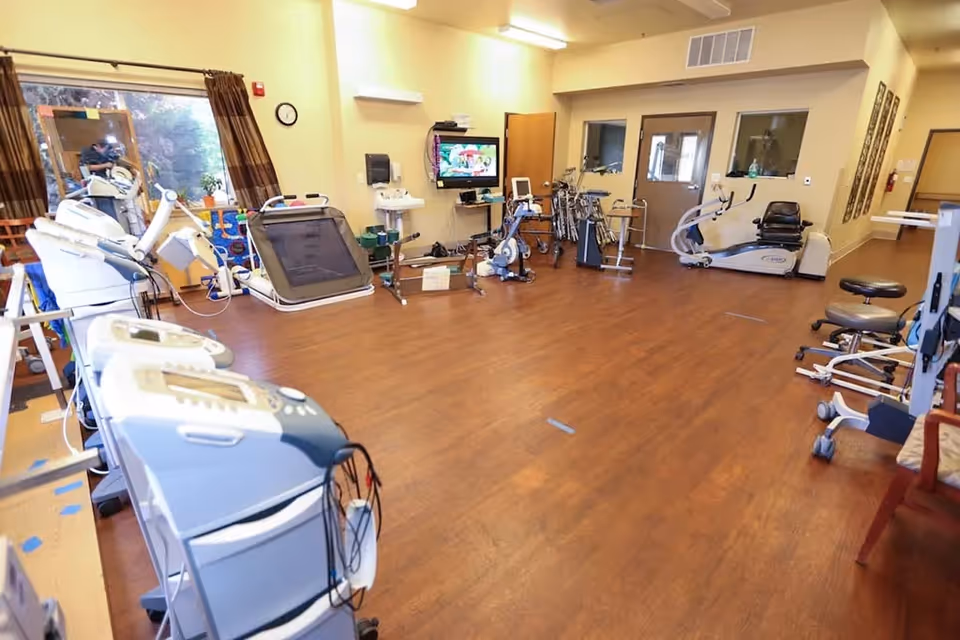 A rehabilitation center room with various exercise and therapy equipment including stationary bikes, treadmills, and other physical therapy machines. The room has wooden flooring, beige walls, a wall-mounted TV, and a large window with brown curtains letting in natural light.