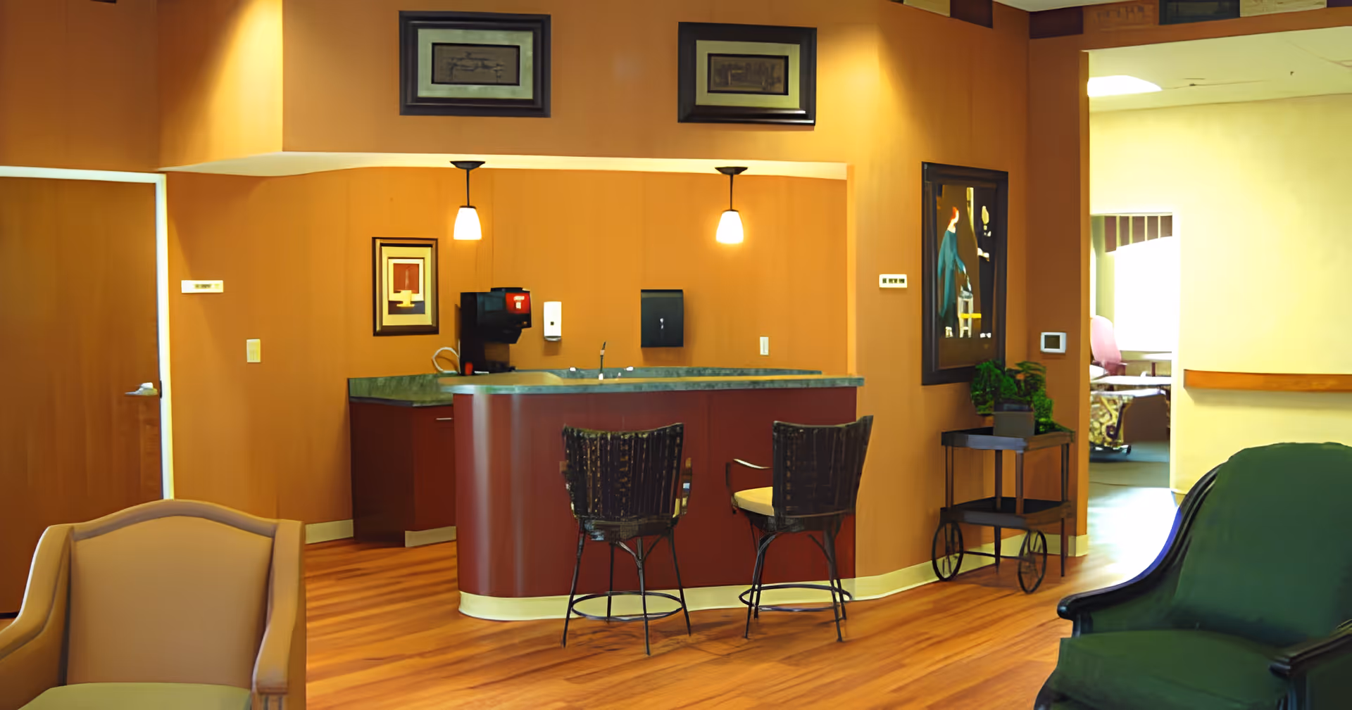 Interior view of a cozy common area in a retirement community featuring a small kitchen counter with two high chairs, pendant lights, framed artwork on the walls, a green armchair, and a beige armchair. The space has warm orange walls and wooden flooring, with a hallway leading to another room in the background.