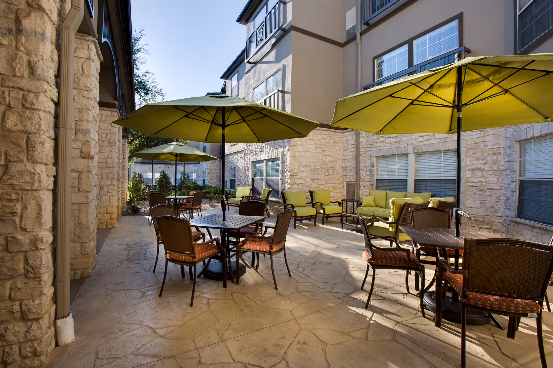 Outdoor patio area at The Reserve at North Dallas Assisted & Senior Living with multiple tables and chairs under large green umbrellas, surrounded by stone walls and windows. There are cushioned seating areas along the wall on the right side.