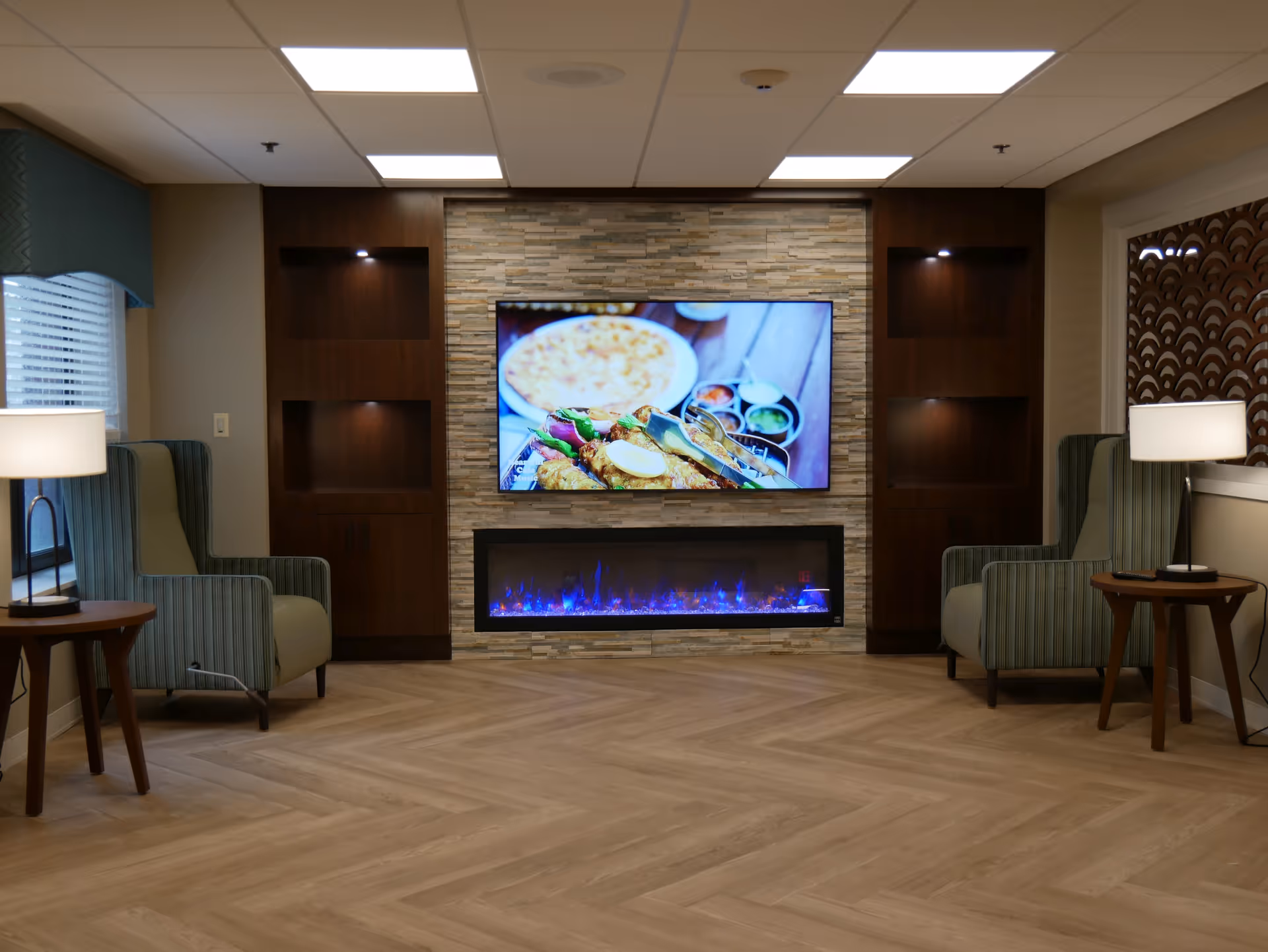 A cozy seating area with two striped armchairs facing a wall-mounted flat screen TV displaying a food image. Below the TV is a modern electric fireplace with blue flames. The wall behind the TV and fireplace is decorated with light-colored stone tiles, flanked by dark wooden built-in shelves with spotlights. Two side tables with lamps are positioned next to each armchair. The room has a light wood herringbone floor and a white ceiling with recessed lighting.