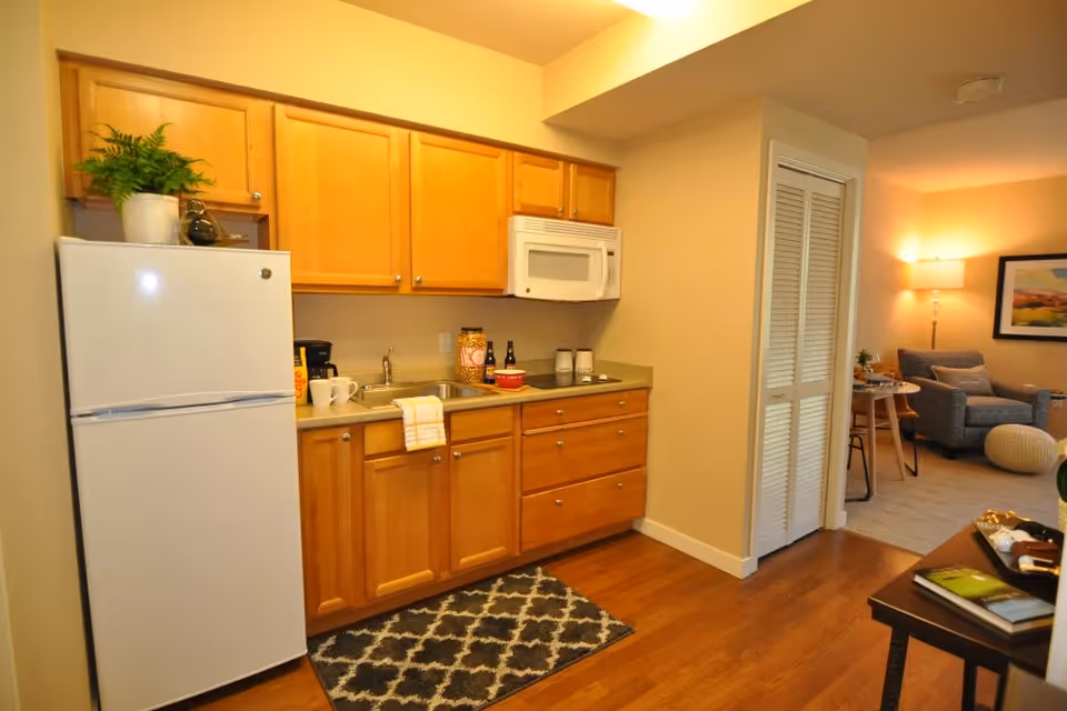 Small kitchen area with wooden cabinets, a white refrigerator and microwave, a sink and countertop, opening into a cozy living area.