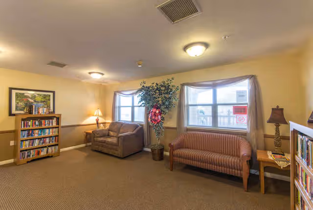 A cozy senior living facility common area with two windows letting in natural light. The room features a brown loveseat, a striped sofa, a tall potted plant with a red decoration, two wooden side tables with lamps, and two bookshelves filled with books. The walls are painted beige with a wooden chair rail, and the ceiling has two light fixtures.